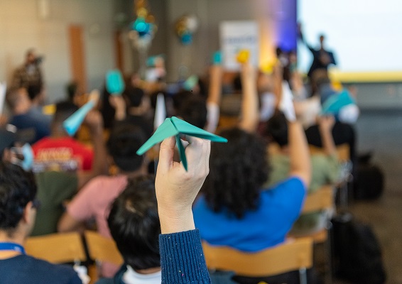 A roomful of people, seen from the back, hold up blue, white and gold paper planes, poised to throw toward an out-of-focus speaker at the front of the room
