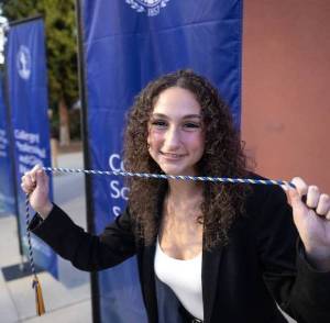Young woman smiling, holding a blue and gold honor cord, standing outside with blue college banners.