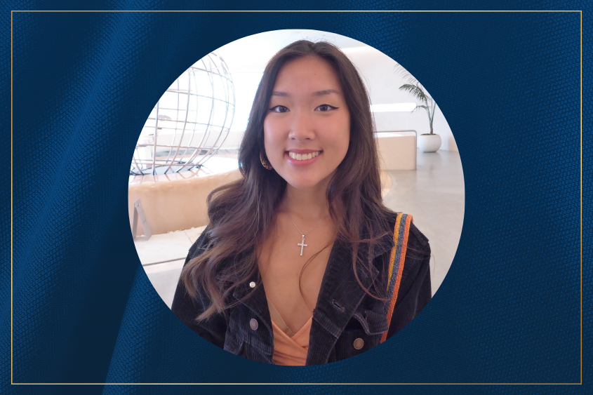 A person with long, wavy hair smiling and wearing a black jacket and a cross necklace stands in a modern, brightly lit space with a large globe decor.
