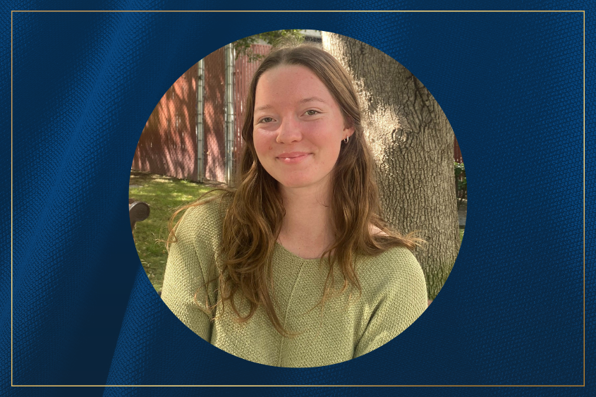 A woman with long, wavy hair wearing a green sweater sits outdoors in front of a tree and a wooden fence.