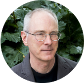 Professor Neil Switz A person wearing glasses and a dark jacket stands among a bush with large green leaves.