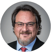 Provost Del Casino A man in a formal black suit and light blue shirt, wearing a red tie with a subtle pattern, poses against a neutral backdrop.